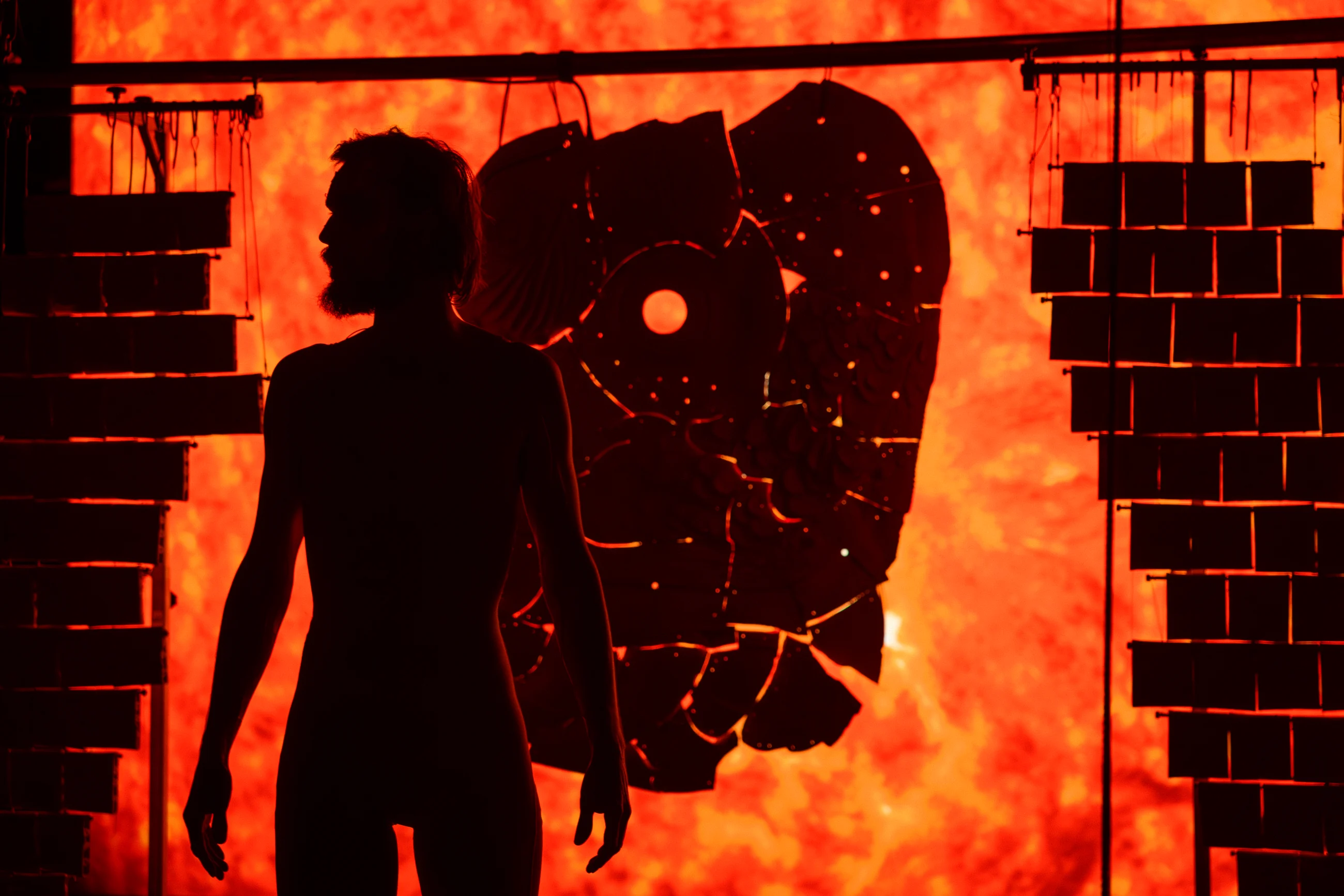 A performer in silhouette before an orange projection and suspended ceramic sculpture.