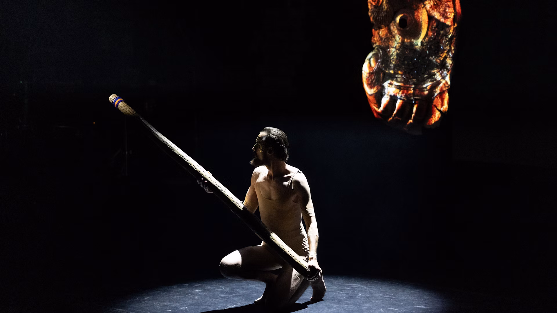A performer kneels with a long ceramic instrument below a suspended form.